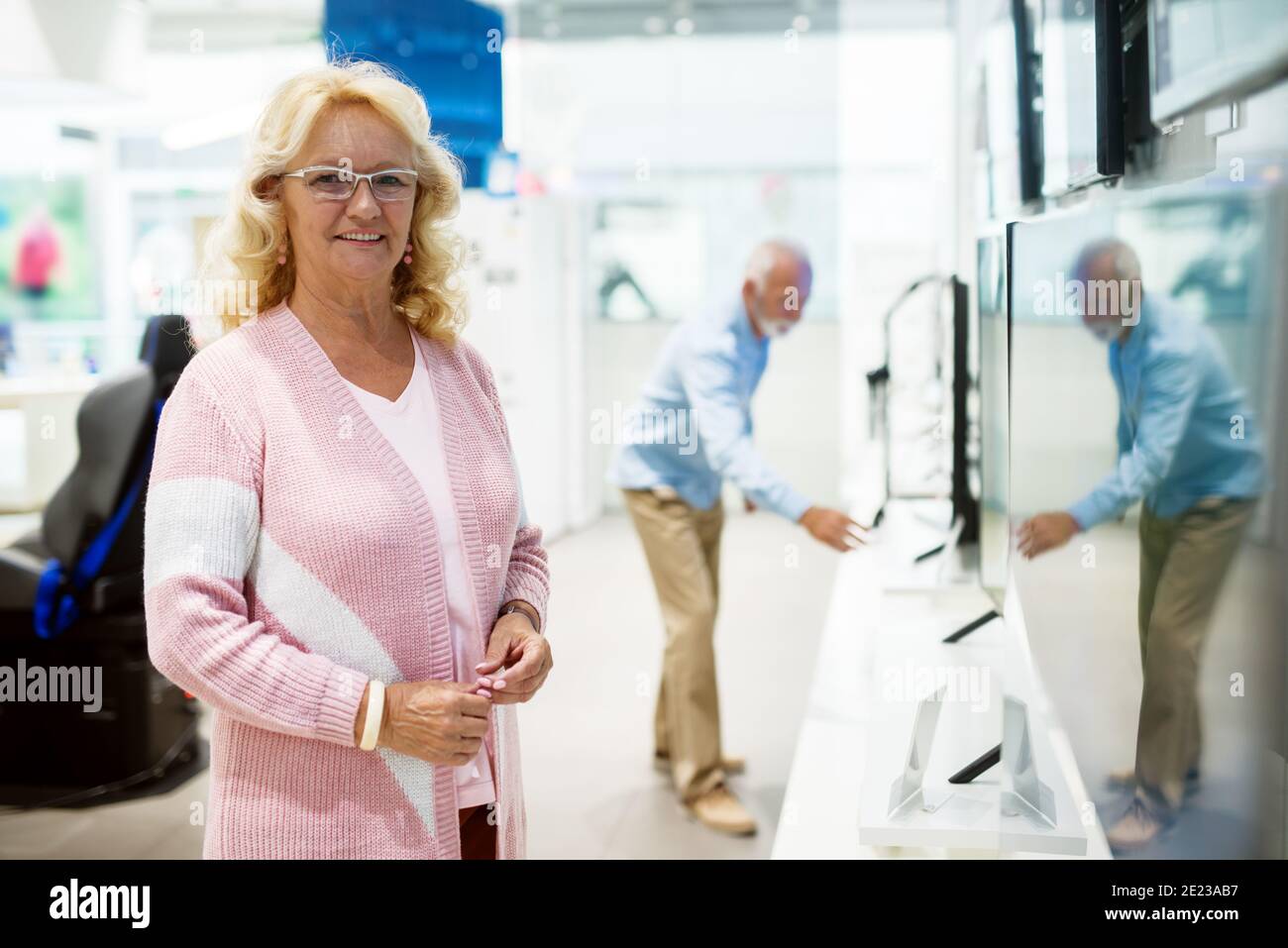 Bella donna caucasica anziana in posa in negozio di tecnologia. In background suo marito in cerca di nuovo televisore. Foto Stock