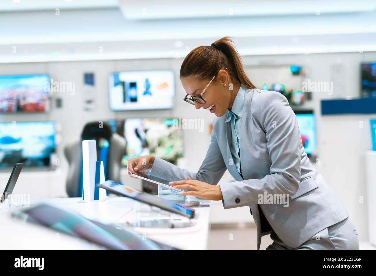 Donna caucasica felice con coda di pony vestita in abiti da lavoro cercando forte nuovo tablet da vicino mentre si sta in piedi vicino al negozio di tecnologia. Foto Stock