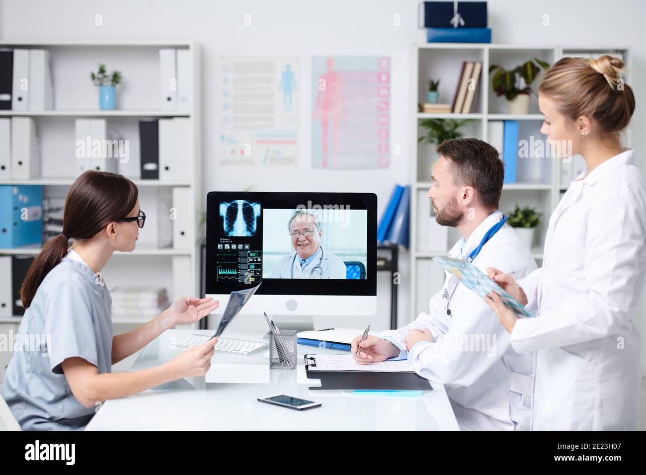 Gruppo di giovani professionisti seduti alla scrivania di fronte monitoraggio del computer e consulenza con il medico senior durante la comunicazione online Foto Stock