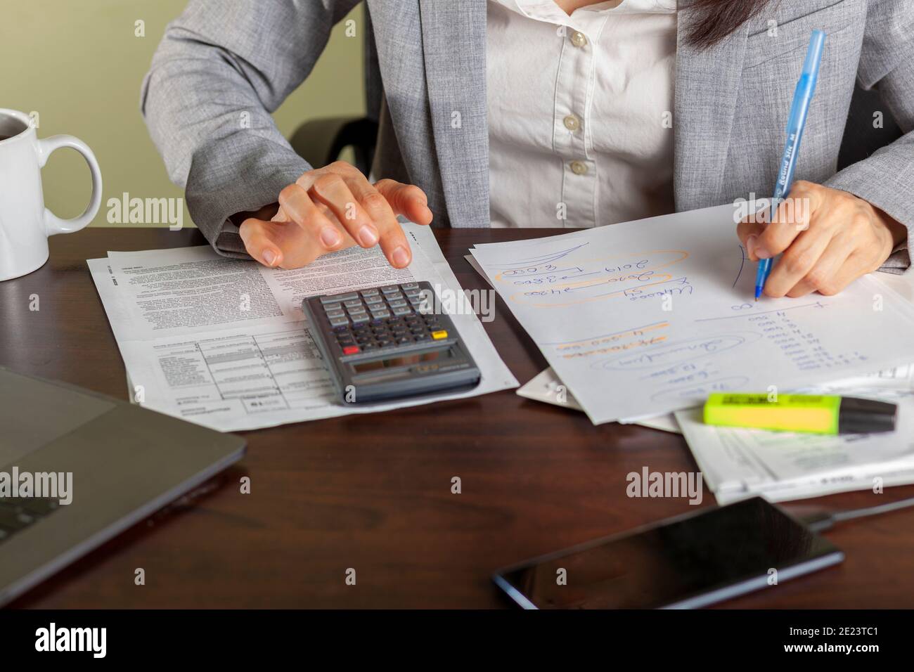 Una donna d'affari che indossa un abito formale sta lavorando in un ambiente d'ufficio. Sta calcolando i redditi e le spese usando gli estratti conto, le polizze di paga, le ricevute ma Foto Stock