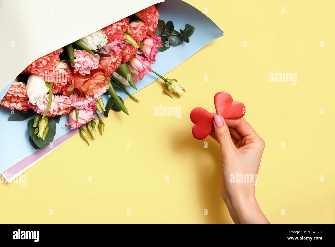 Vista dall'alto delle mani femminili con due cuori e un bouquet di fiori su sfondo giallo. Concetto di San Valentino, simbolo di amore, romanticismo. Foto Stock