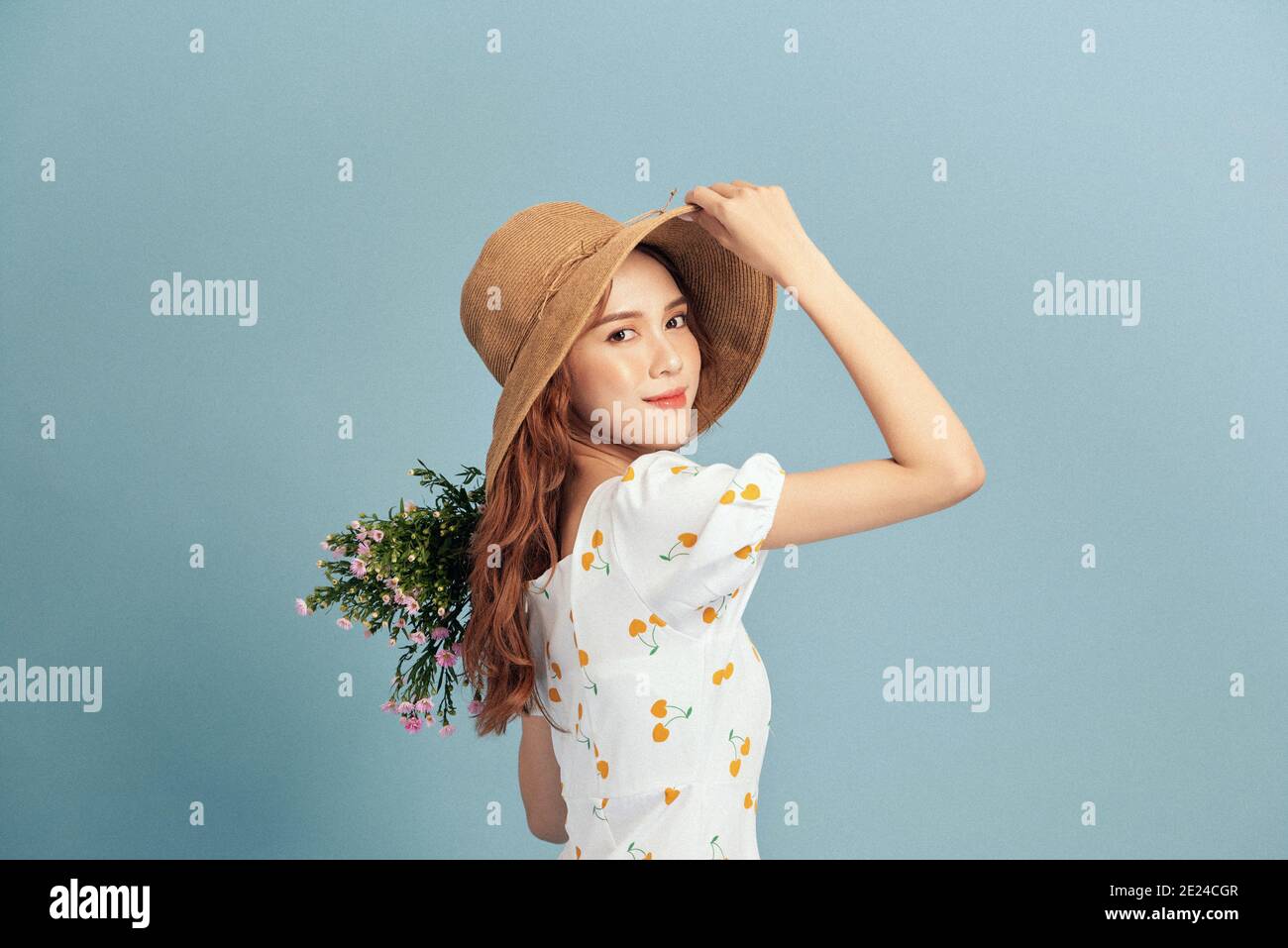 Primo piano ritratto di una giovane donna attraente in estate abito e cappello di paglia che tiene bouquet di fiori e guardando sopra la sua spalla isolata su blu b Foto Stock