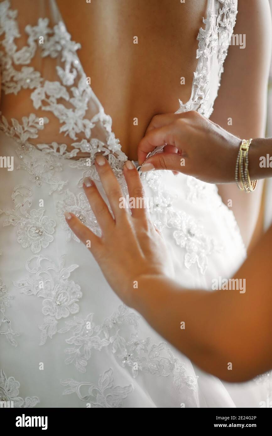 Donna che assiste una sposa a vestirsi nel suo elegante bianco abito con rifiniture in pizzo in preparazione alla cerimonia nuziale in primo piano sulle sue mani Foto Stock