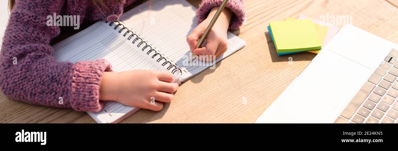 Vista ritagliata della ragazza che scrive in notebook vicino a note appiccicose e laptop sulla scrivania, banner Foto Stock