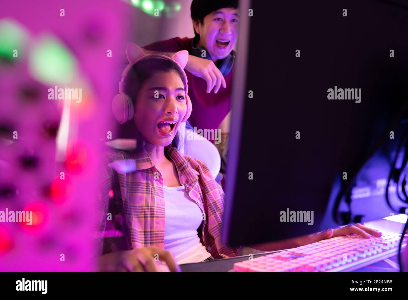 Giovane attraente donna asiatica che gioca a videogiochi nel suo studio con il suo ragazzo sul retro. Vista laterale di una coppia che lavora di fronte alla stessa workstation in uno studio di design professionale Foto Stock