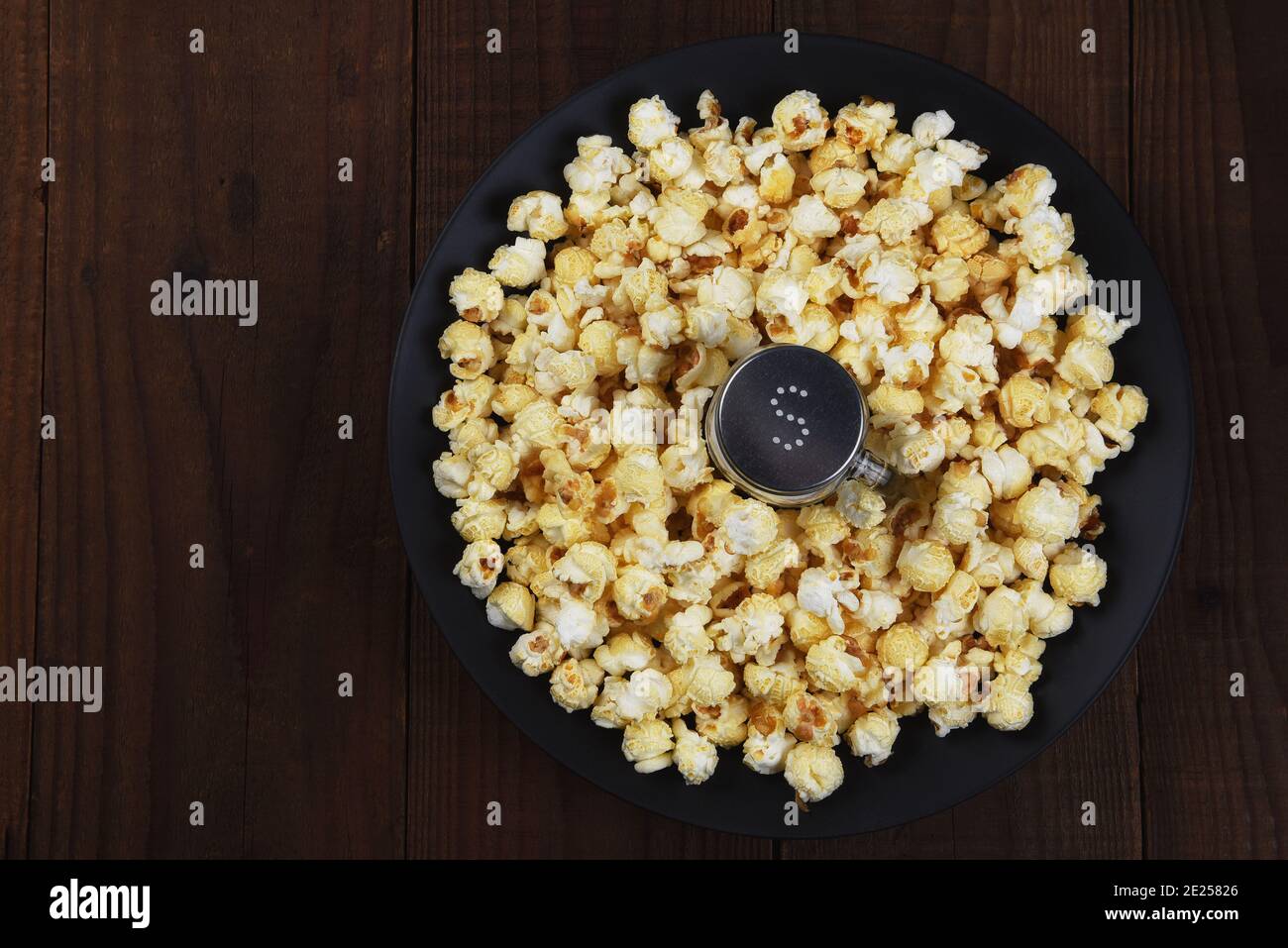 Colpo in testa di uno shaker di sale in una ciotola poco profonda circondata da popcorn appena fatto. Formato orizzontale su tavola scura rustica con spazio per la copia. Foto Stock