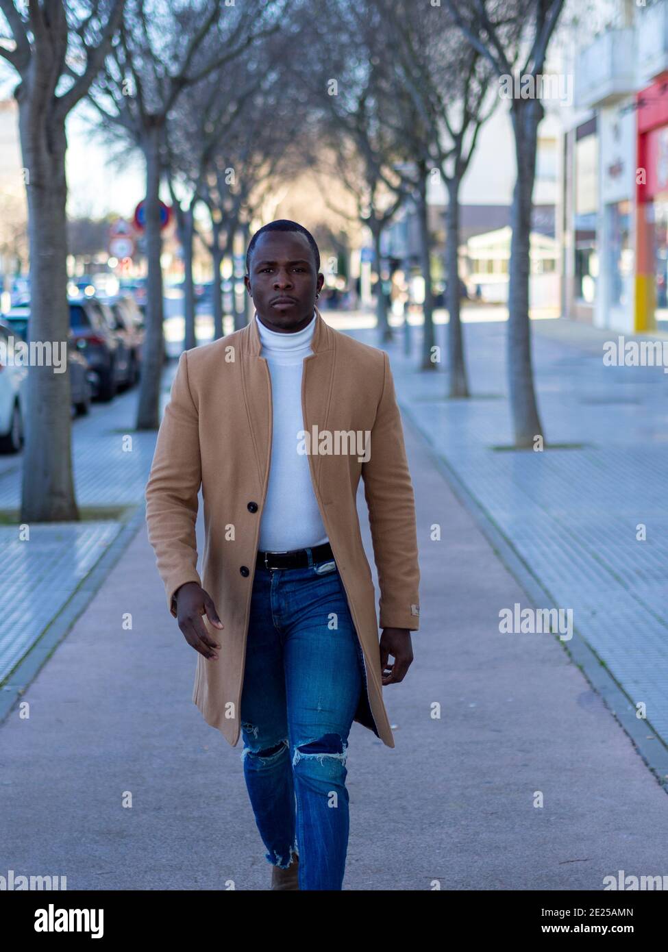 Elegante uomo africano che indossa jeans con un maglione bianco e. cappotto  beige mentre camminando giù per la strada Foto stock - Alamy