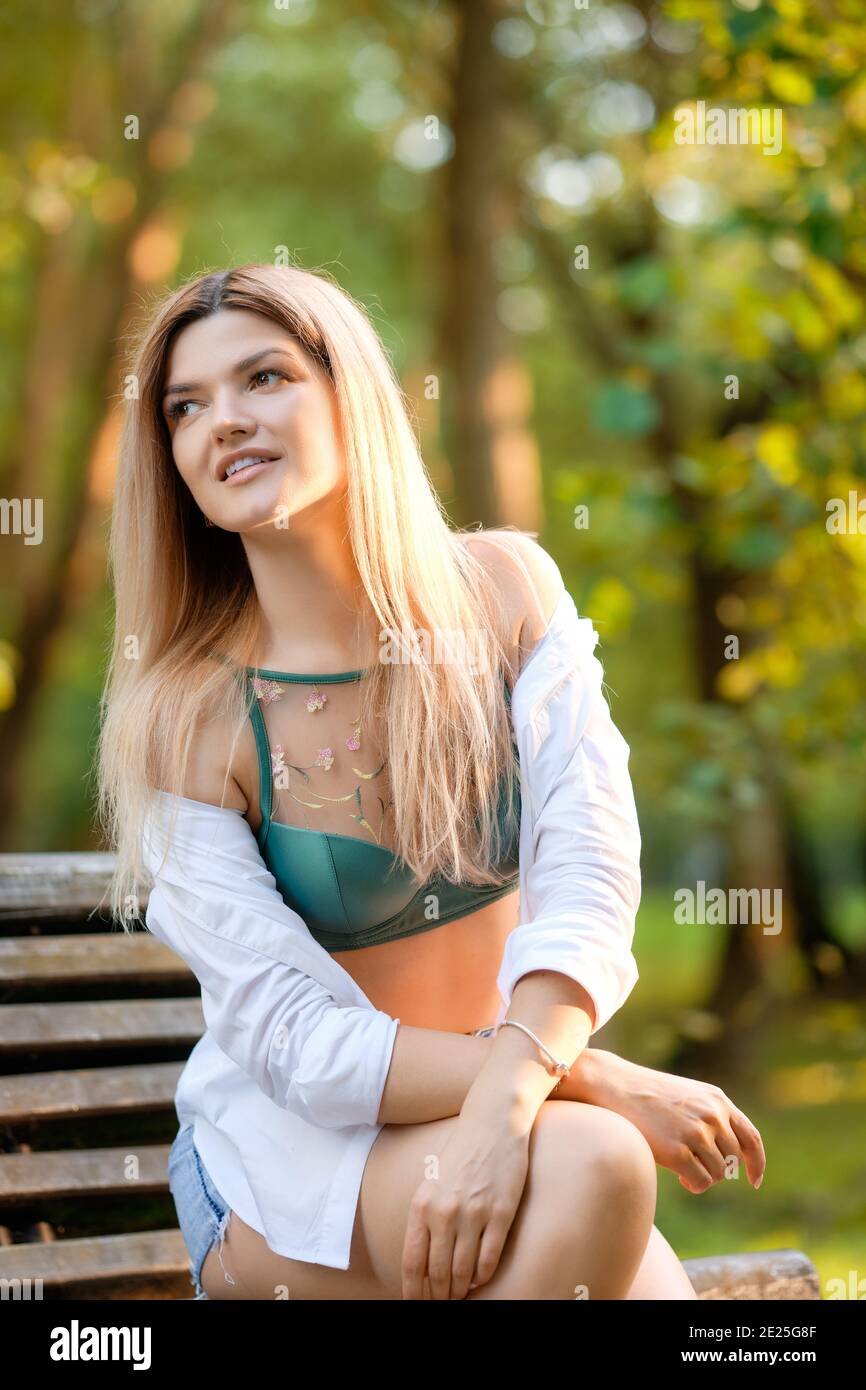 Carina ragazza seduta alla panchina sotto la luce del sole nel parco estivo. Indossa pantaloncini jeans e camicia bianca allacciata in vita. Foto Stock