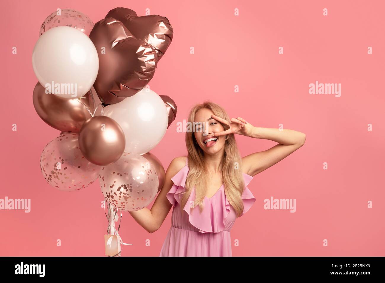 Sciocco giovane donna con palloncini di compleanno che mostra la sua lingua, winking e gesturing pace su sfondo rosa studio Foto Stock