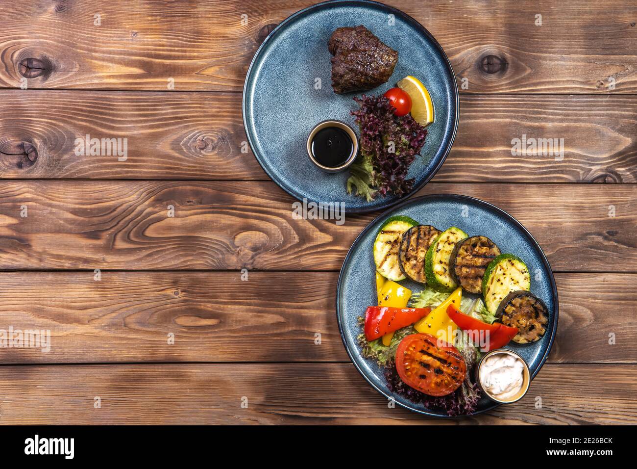 Bistecca di manzo con limone, pomodoro, lattuga e verdure grigliate su piatti su fondo di legno marrone. Layout piatto. Foto Stock