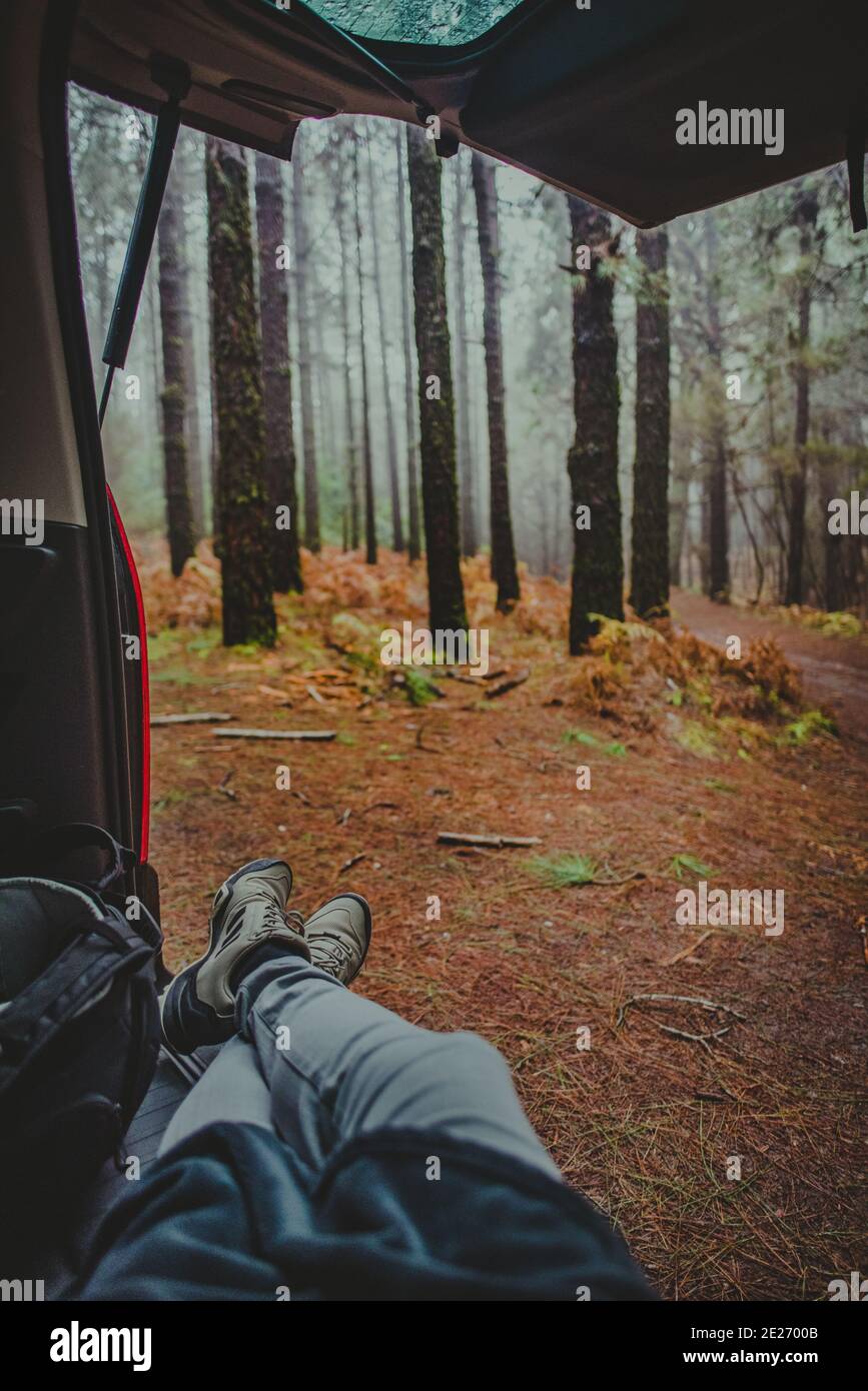 I piedi di una persona sono visti poggiare all'interno di un veicolo di camper nel mezzo del campo, tronchi di alberi grandi sono visti sullo sfondo Foto Stock