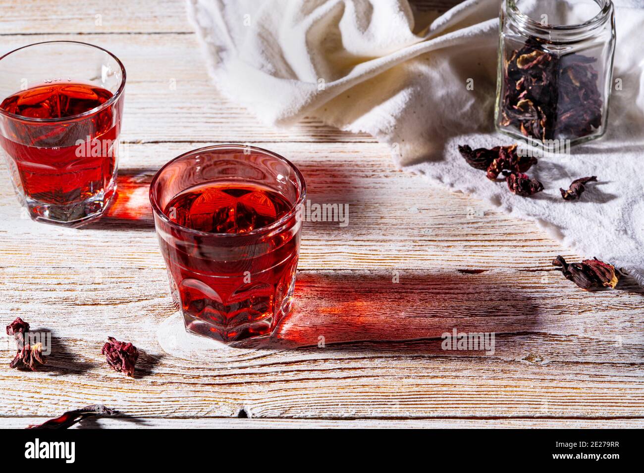 Rinfrescante tè hibiscus in bicchieri su tavolo di legno, petali di rosa e tè secco su tavolo di legno bianco rustico. Luce dura con ombre dure Foto Stock