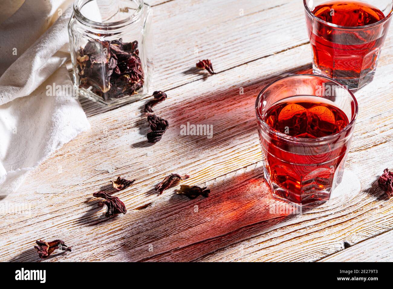 Rinfrescante tè hibiscus in bicchieri su tavolo di legno, petali di rosa e tè secco su tavolo di legno bianco rustico. Luce dura con ombre dure Foto Stock