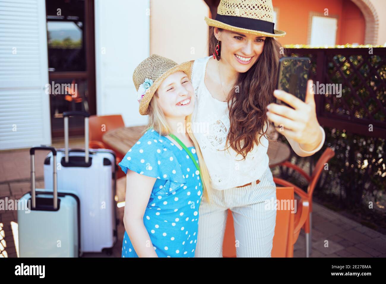 sorridente madre e figlia trendy in cappelli con borse ruota che hanno videochiamata su uno smartphone sulla terrazza dell'hotel della pensione. Foto Stock