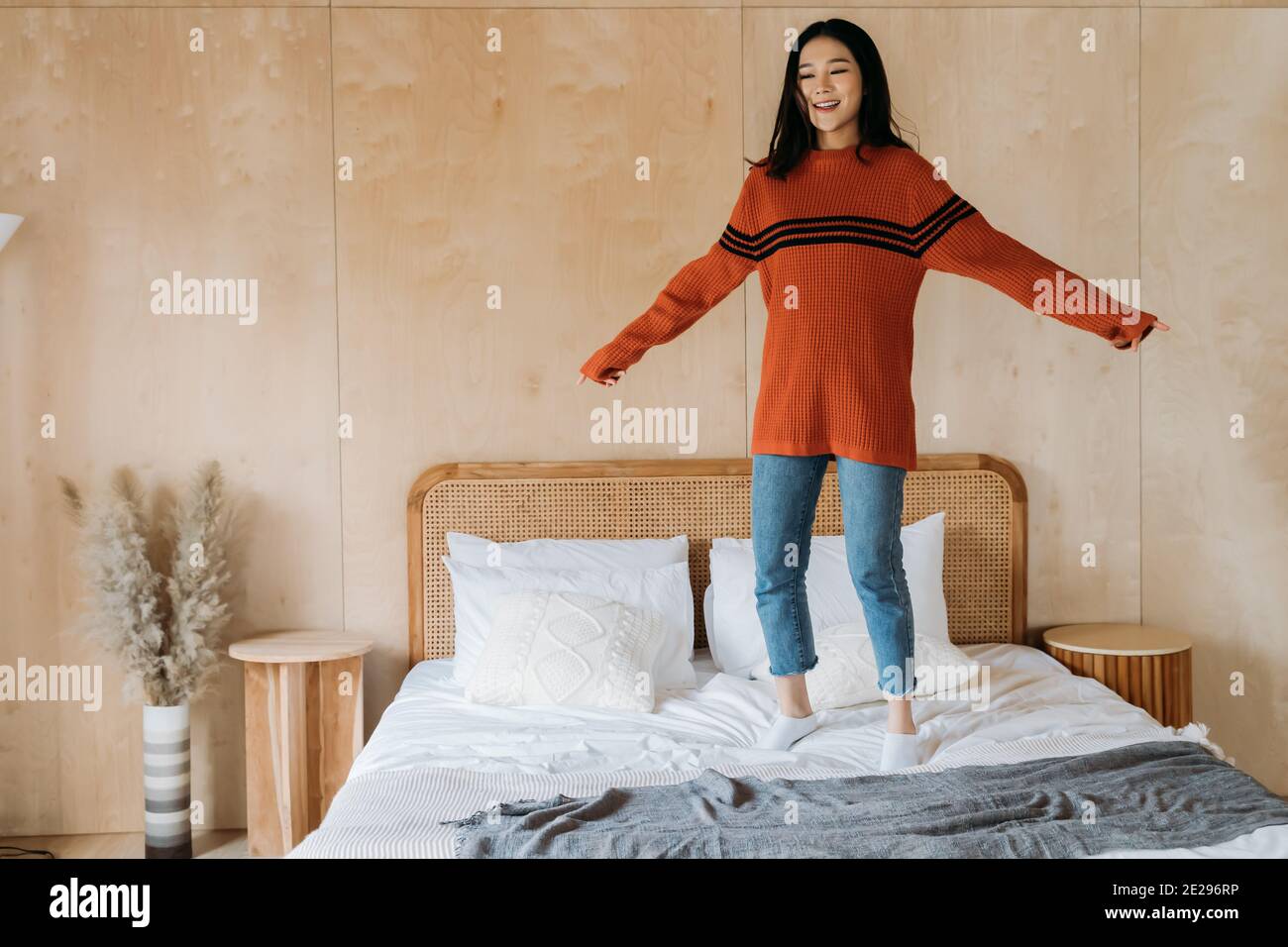 Allegra e rilassante donna etnica asiatica in maglione sorridente e saltellante a letto mentre ci si diverte a casa Foto Stock