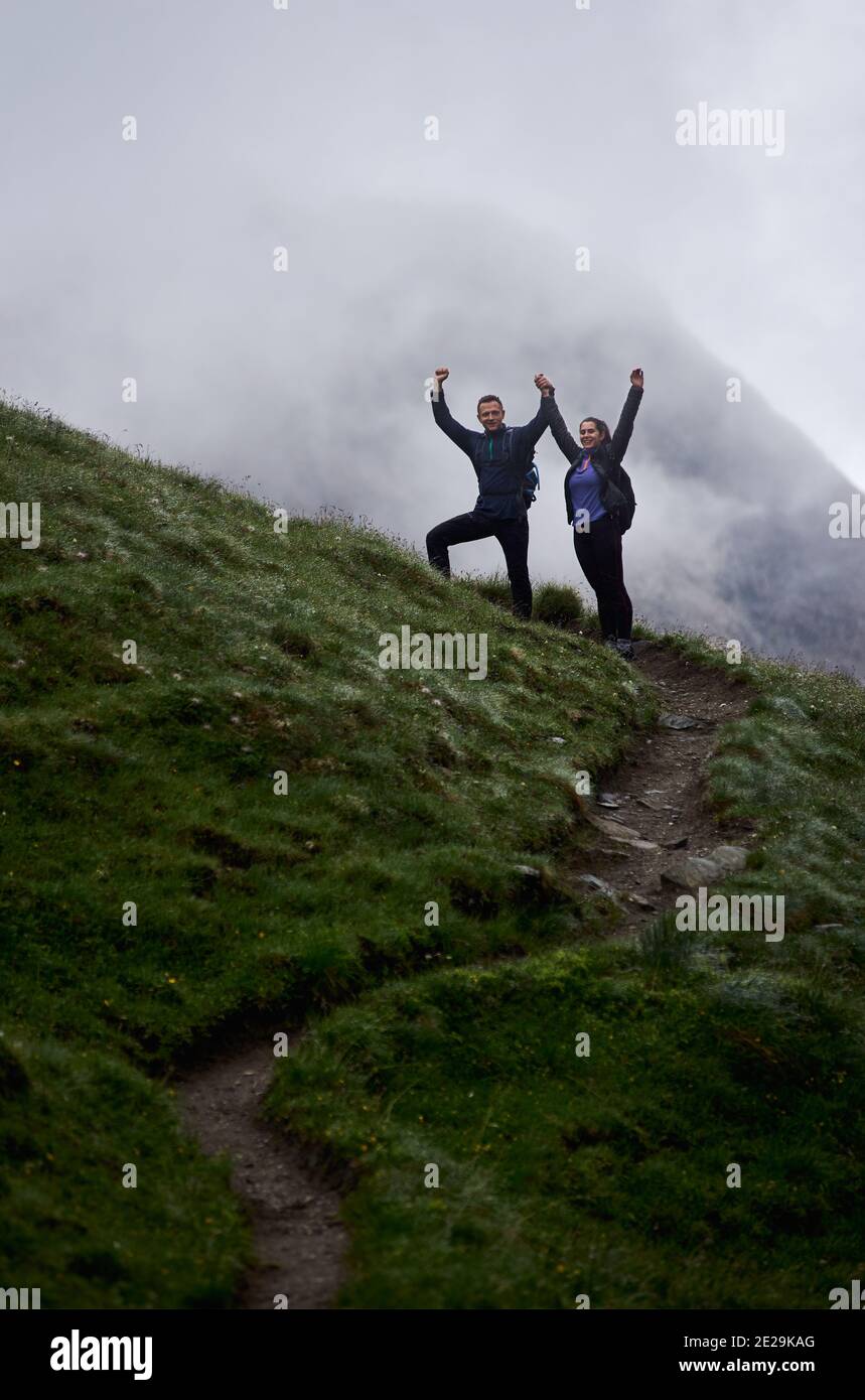 Tutta la lunghezza di due viaggiatori che si trovano su un sentiero in collina e alzano le braccia. Giovane donna e ragazzo che tiene le mani e si diverte a fare un'escursione in montagna. Concetto di escursioni, viaggi e gioia. Foto Stock