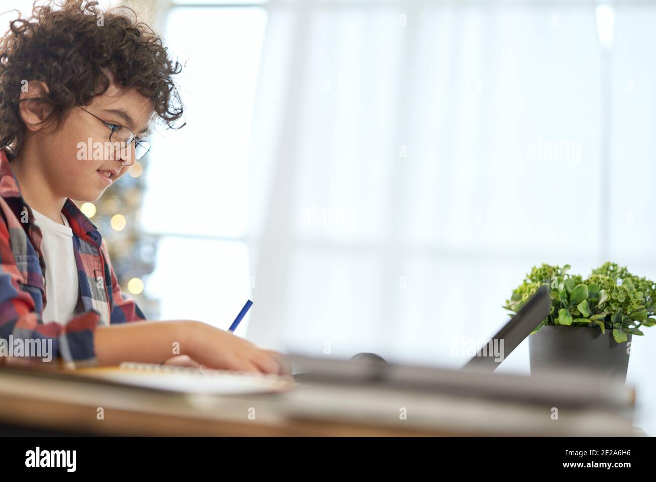 Entusiasta ragazzo della scuola latina che indossa occhiali che fanno i compiti con un tablet digitale, seduto alla scrivania mentre studia a casa. Formazione online, tecnologia, homeschooling. Vista laterale Foto Stock