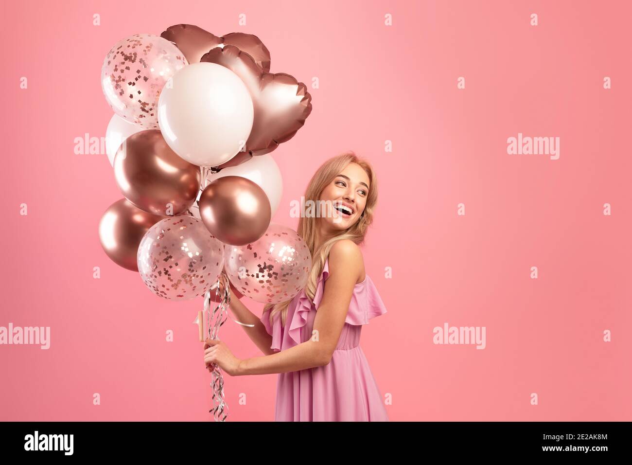 Tempo di festa. Affascinante donna bionda in abito elegante che tiene palloncini di compleanno su sfondo rosa studio, spazio di copia Foto Stock