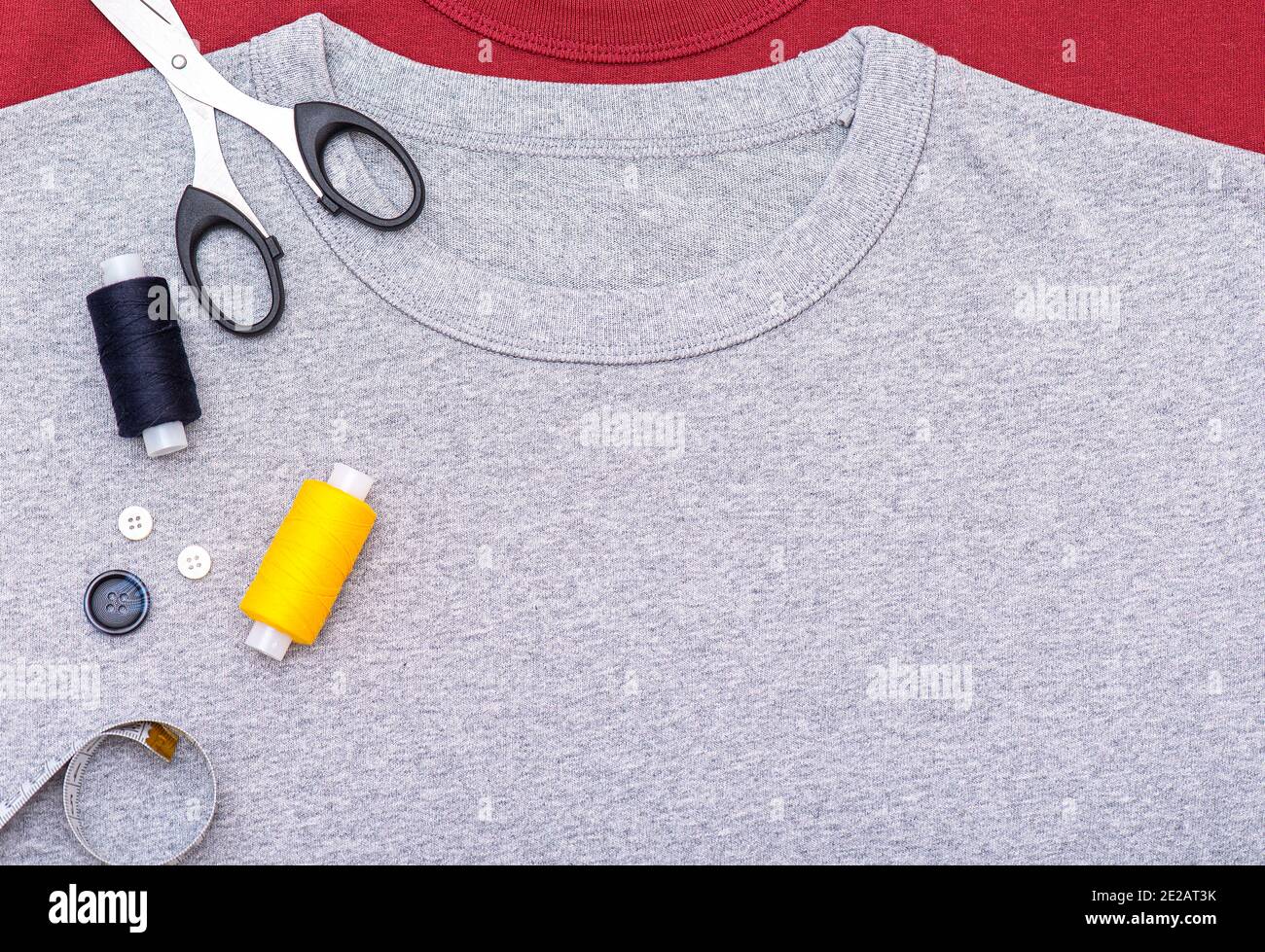Composizione semplice con bottoni, metro a nastro, forbici, bobina di filo. Attrezzatura da cucire su t-shirt grigia, sdraiatura, vista dall'alto. Foto Stock