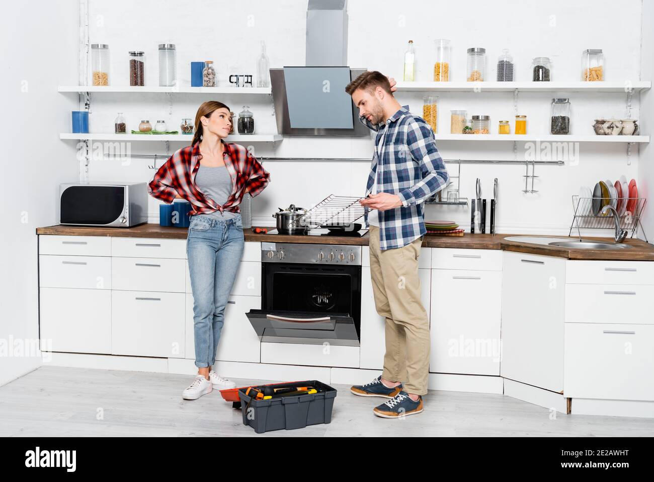 tutta la lunghezza della donna insoddisfatta che guarda l'uomo che tiene la griglia vicino a forno disassemblato e toolkit in cucina Foto Stock