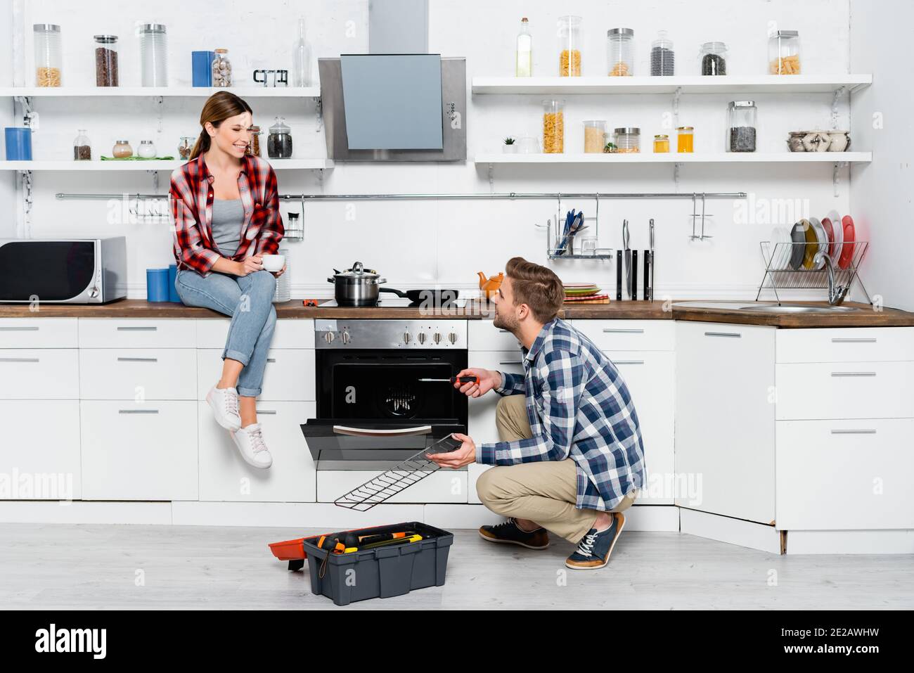 lunghezza intera di donna sorridente con tazza di caffè guardando all'uomo con griglia vicino al forno e toolkit in cucina Foto Stock