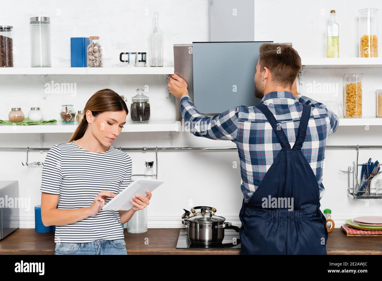 giovane donna che usa la tavoletta vicino a un manipolo che ripara la ventola dell'estrattore dentro cucina Foto Stock