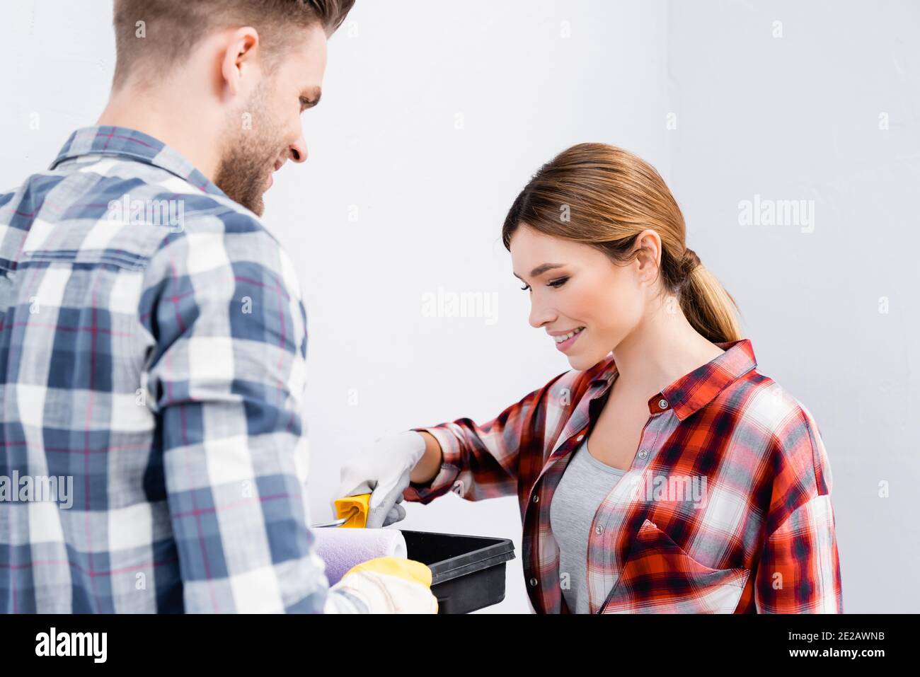 felice giovane donna che mette rullo di vernice in vassoio vicino sfocato uomo in primo piano in interni Foto Stock