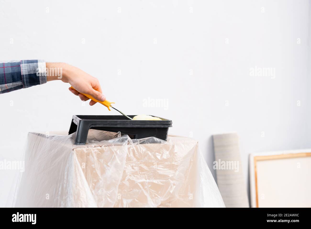 vista ritagliata di un uomo che mette il rullo di vernice nel vassoio scatola coperta con polietilene su sfondo sfocato all'interno Foto Stock