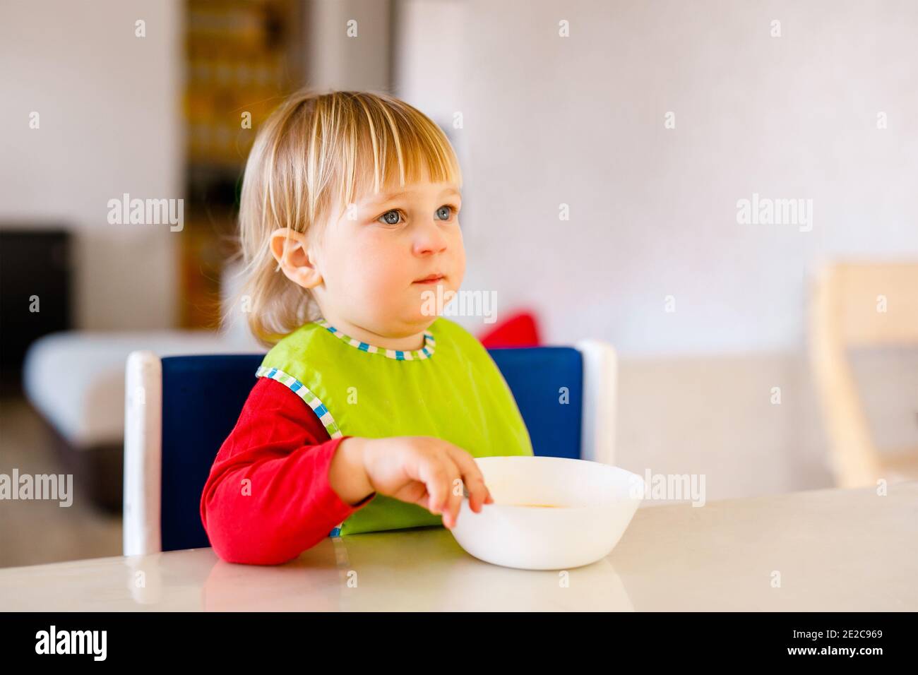 Carino bambino 1,4 anni seduto su seggiolone bambini e mangiare verdure da solo in cucina bianca. Foto Stock