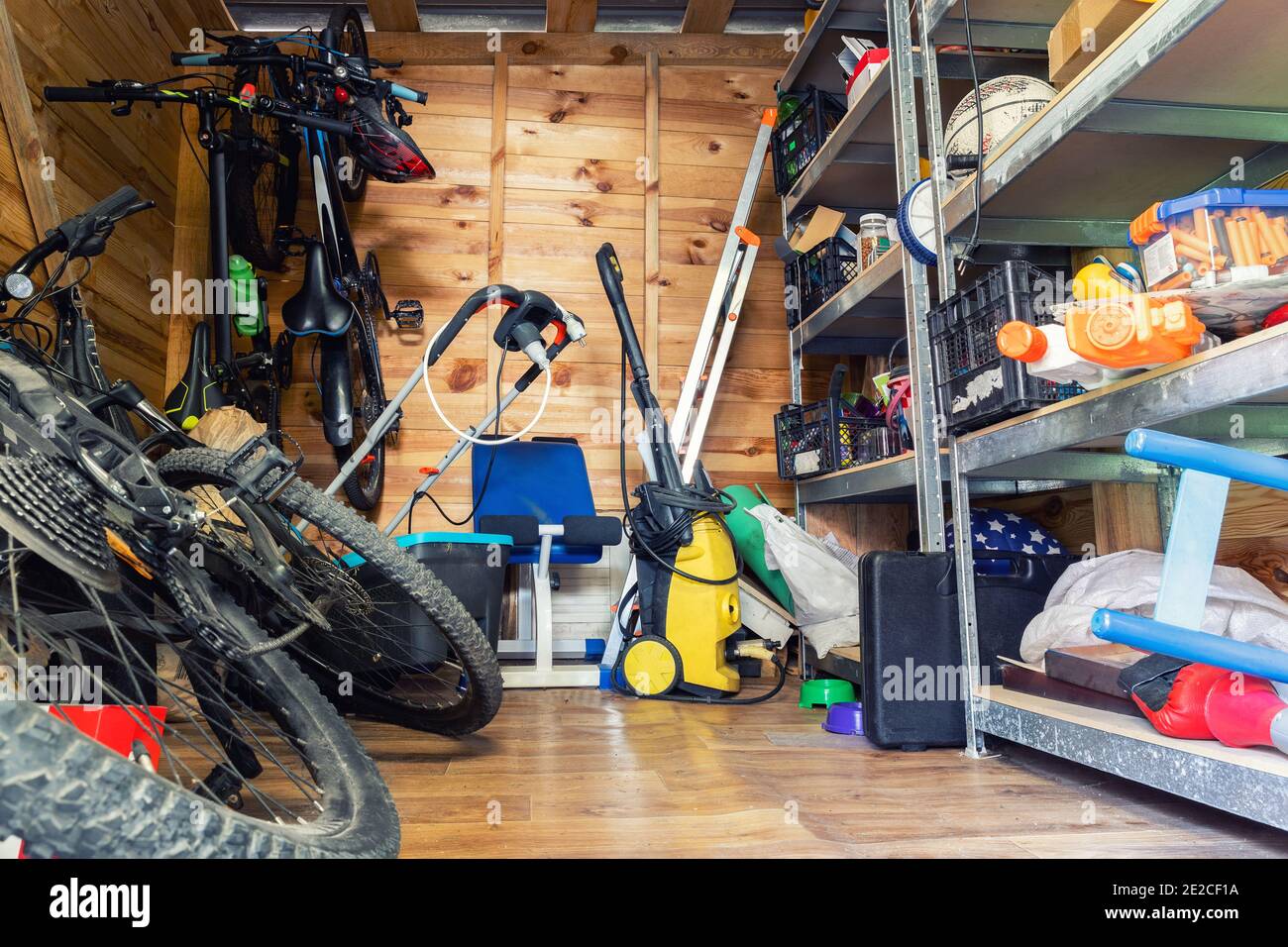 Unità di deposito di utilità di legno della casa suburbana con roba vario su mensole, biciclette, macchina di esercitazione, scala, attrezzi di giardino e attrezzatura. Disordinato Foto Stock