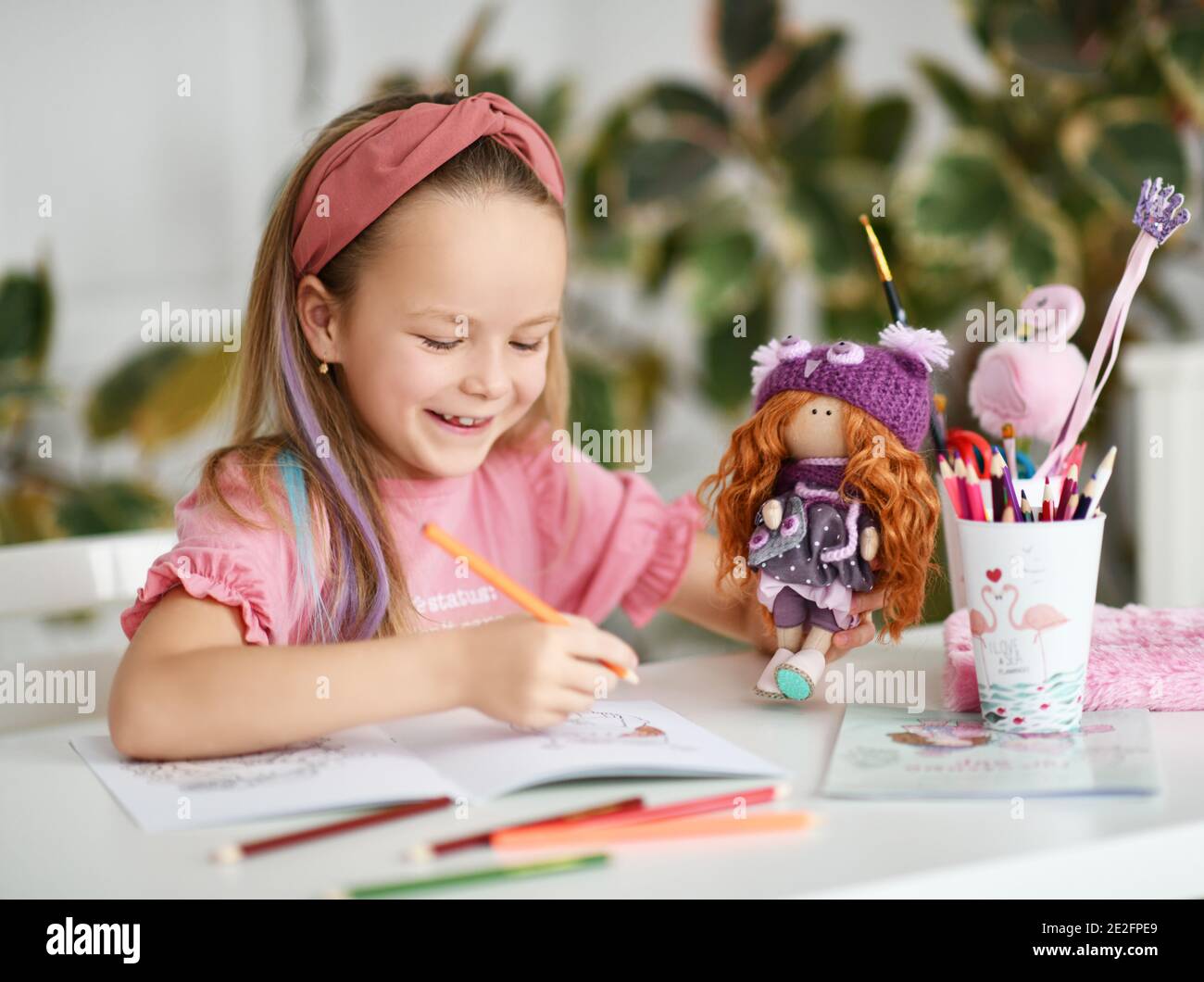 Sorridente piccola bella ragazza con accessorio decorativo per i capelli seduti e colorare il disegno di bambola con matite colorate Foto Stock