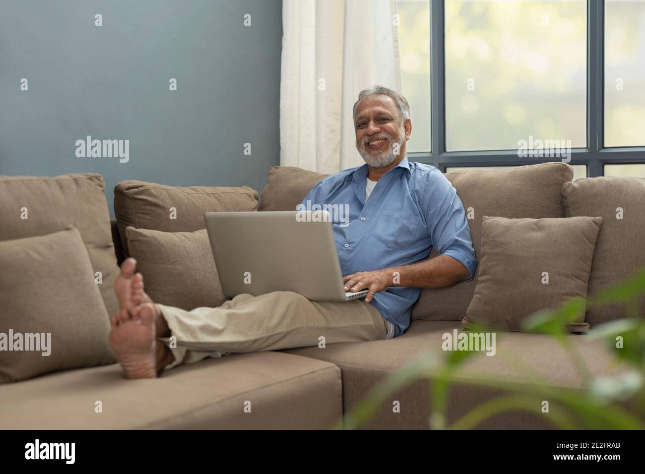 UN VECCHIO FELICE SEDUTO COMODAMENTE SU DIVANO CON COMPUTER PORTATILE Foto Stock