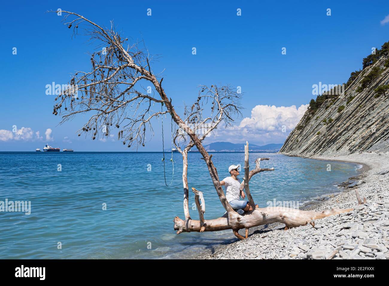 Pittoresco paesaggio estivo su una spiaggia di pietra. Vista mare, un ragazzo seduto su un albero caduto sulla riva, navi da carico e ripide scogliere. Foto Stock