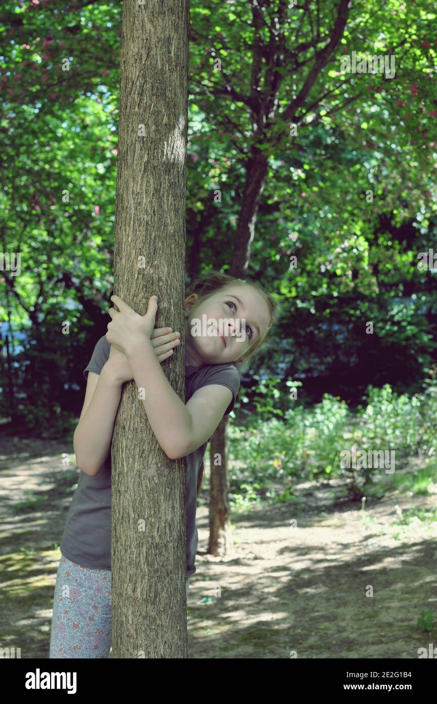 Bambino biondo, ragazza, 8 anni, guardando in su, in piedi dietro un albero e abbracciandolo nella foresta, vista frontale, fuoco selettivo Foto Stock