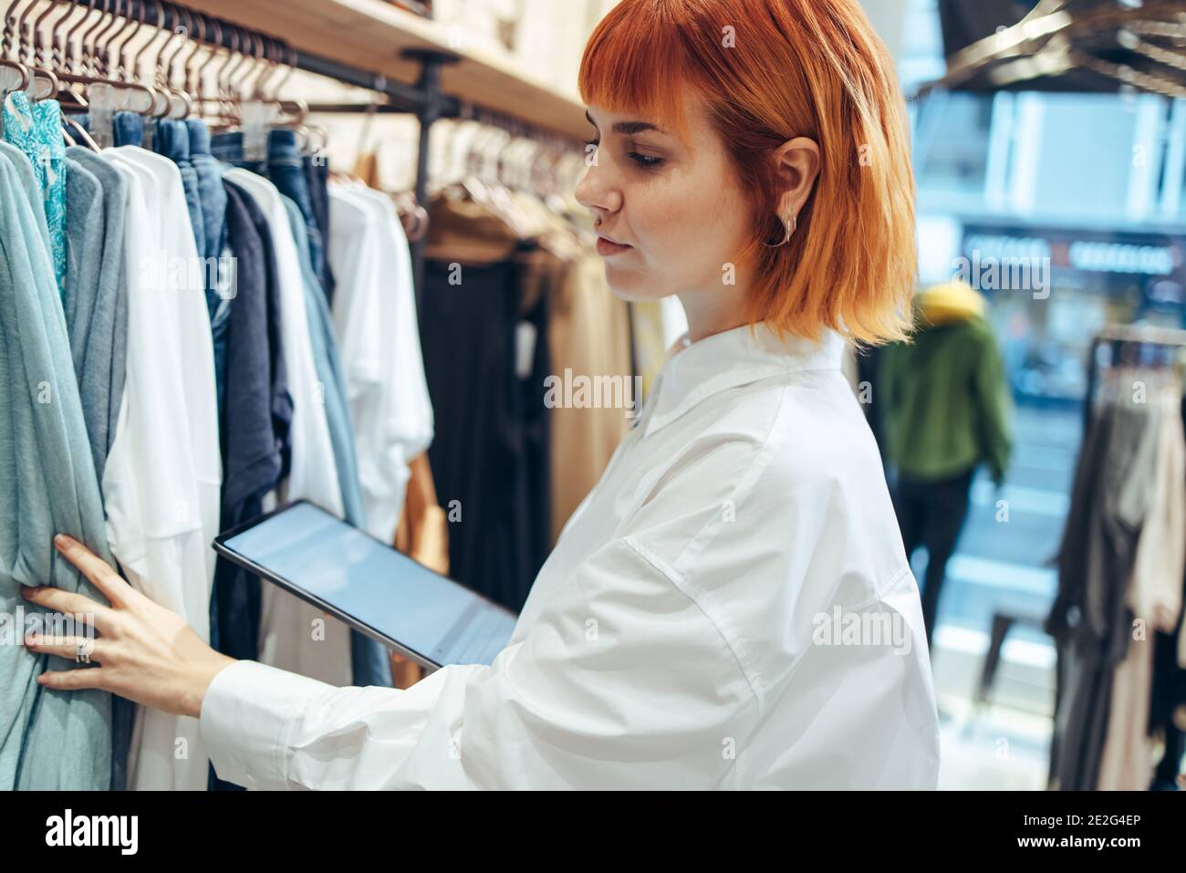 Donna con un tablet digitale che guarda i vestiti appesi in negozio. donna d'affari che lavora in un negozio di abbigliamento che fa il punto sui vestiti. Foto Stock