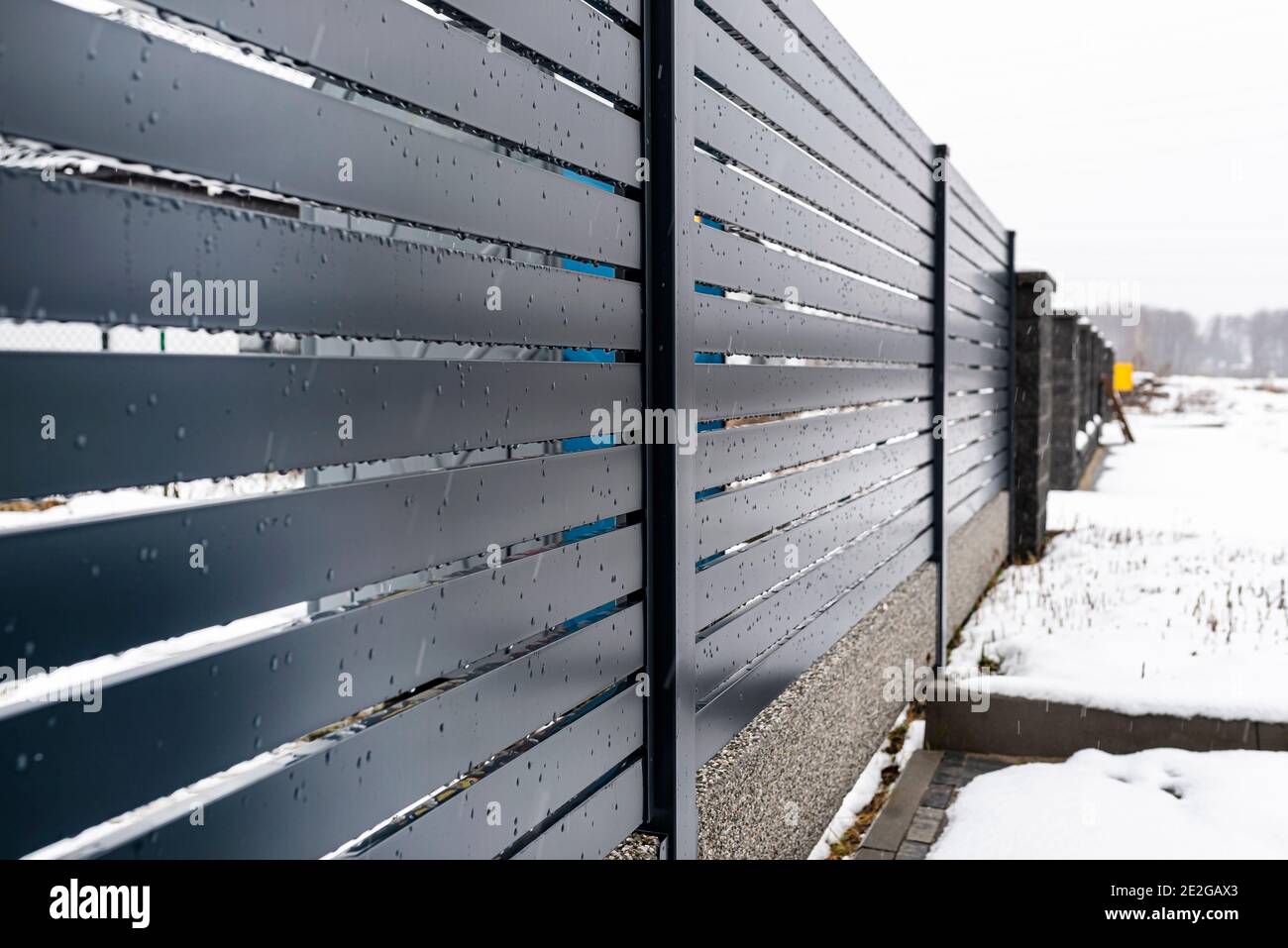 Moderna recinzione antracite, con un connettore di fondazione recinzione visibile, piove in inverno, sullo sfondo c'è neve che giace a terra. Foto Stock