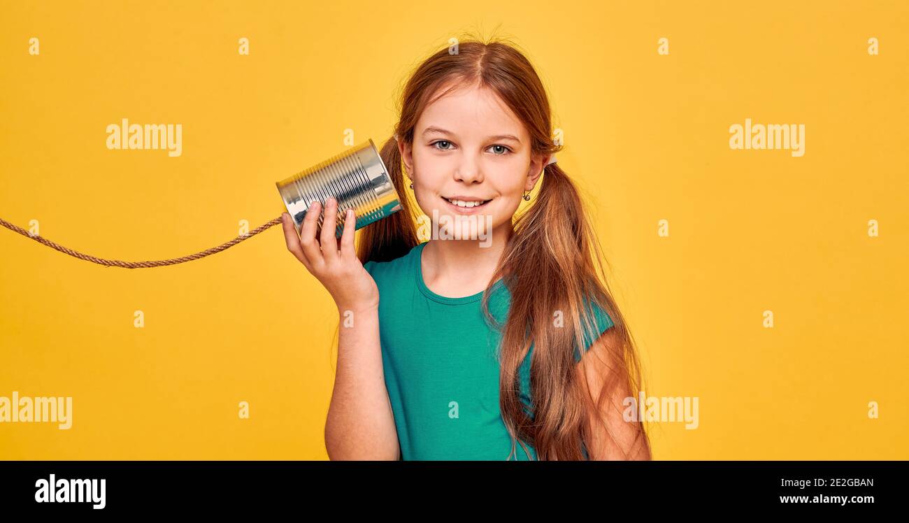 Sorridente ragazza adolescente che ascolta Tin può telefonare, giocando in chat retro connesso, su sfondo giallo. Concetto di cavi incrociati e connessione telefonica Foto Stock