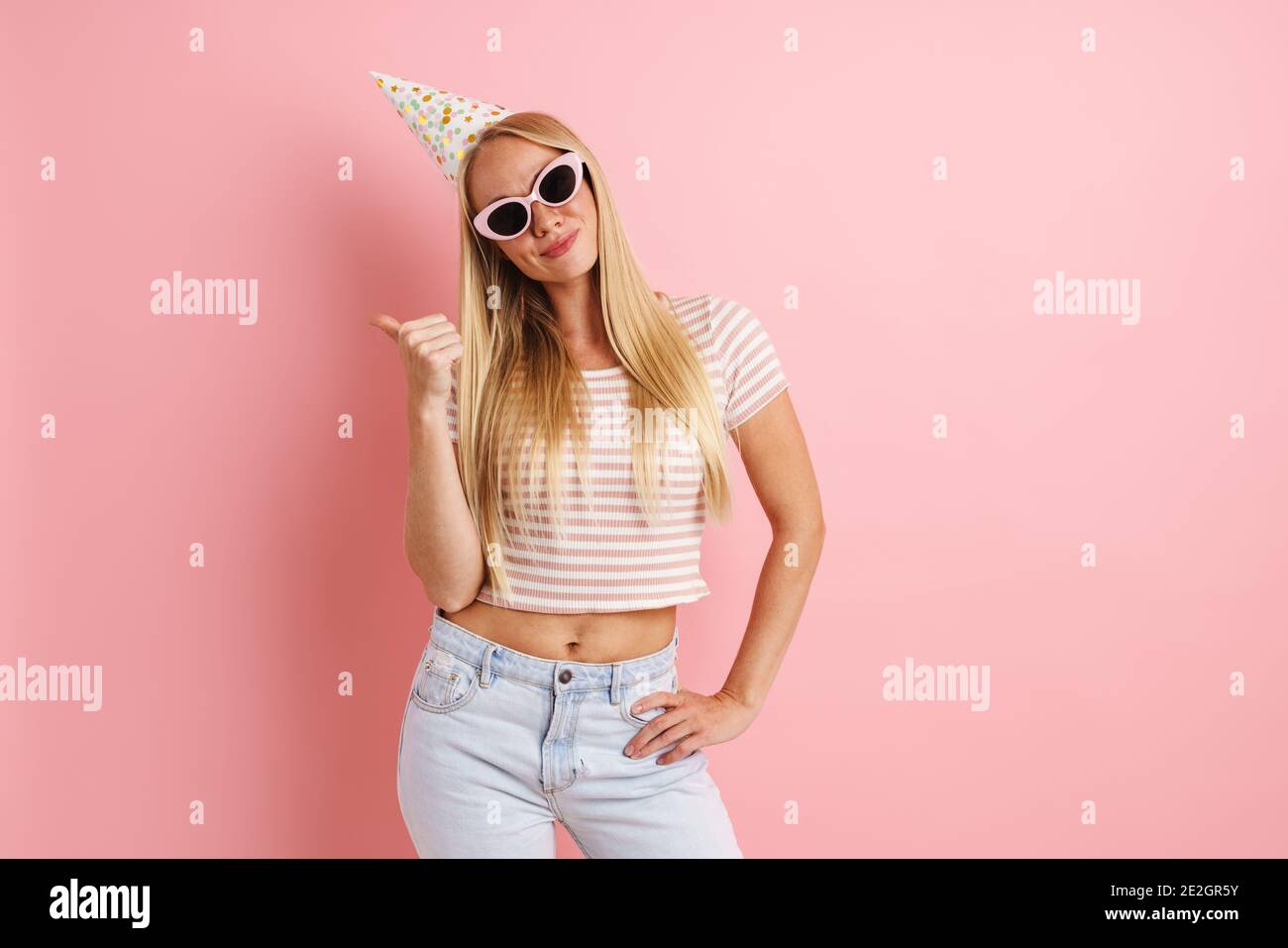 Caucasica gioiosa ragazza in cono di partito sorridente e puntante dito da parte isolato su sfondo rosa Foto Stock