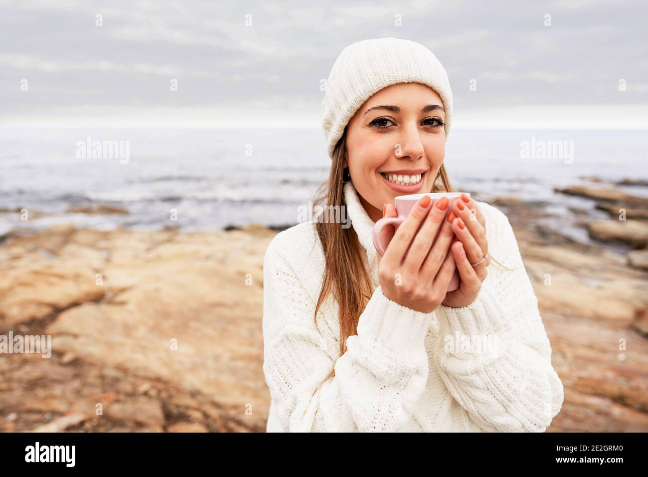 Bella giovane caucasica fiduciosa donna guardando la macchina fotografica con toothy sorridi indossando cappello di lana bianca e maglione che tiene una tazza nelle sue mani all'aperto a. Foto Stock