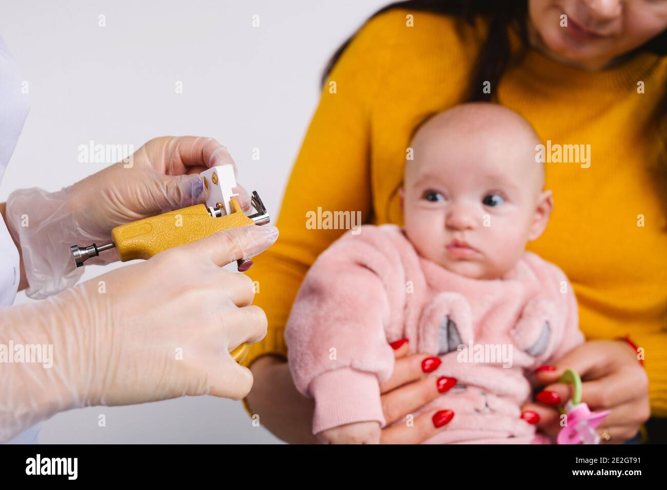Mani del medico in guanti con pistola di piercing e orecchini medici. Sullo  sfondo, mamma con babygirl su sfondo grigio Foto stock - Alamy
