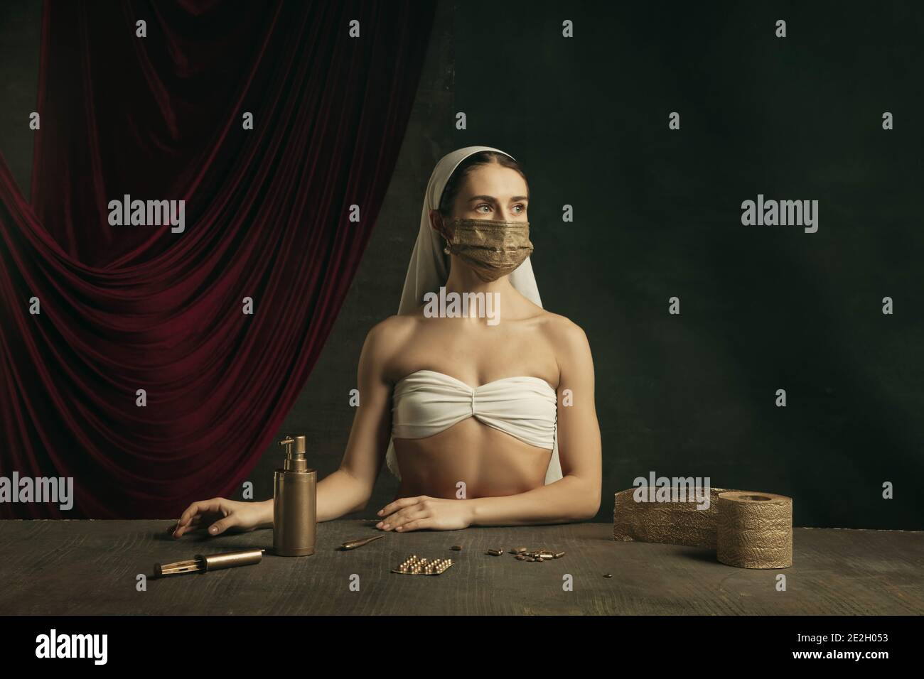 Assistenza sanitaria. Moderno remake di opere d'arte classiche con tema coronavirus - giovane donna medievale su sfondo scuro in posa con maschera dorata e pillole. Concetto di coronavirus, pandemia, creatività. Foto Stock