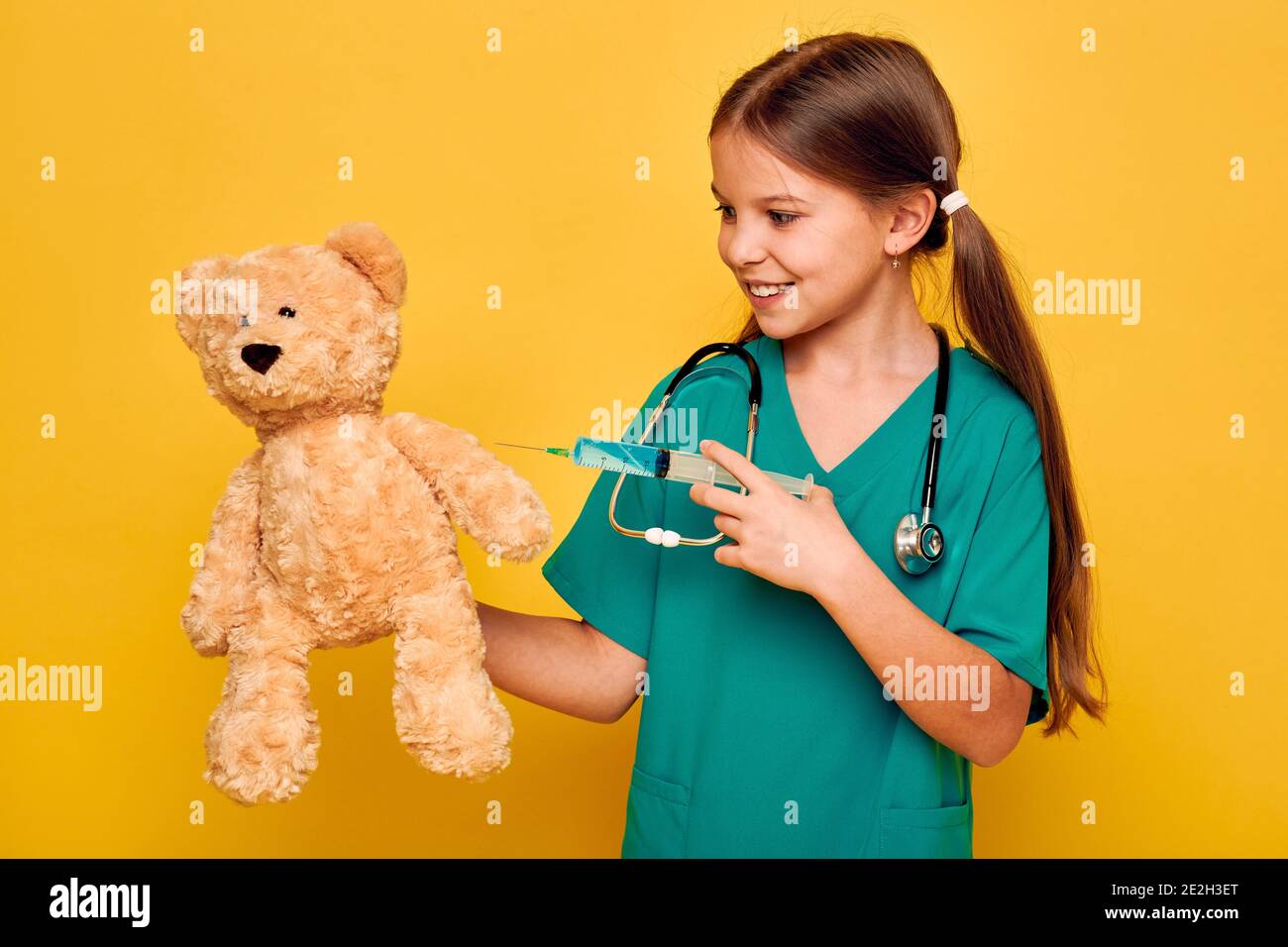 Carina bambina che va iniezione al suo paziente giocattolo orso. Bambino che gioca medico, professione futura. Sfondo giallo Foto Stock