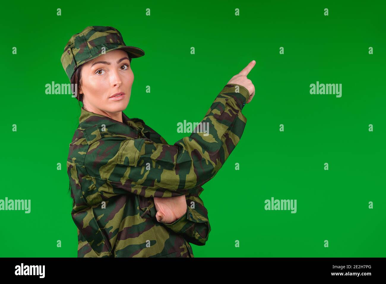 Giovane donna caucasica in un uniforme militare guarda la macchina fotografica e punta con un indice a vuoto spazio laterale. Sfondo verde con spazio laterale. Foto Stock