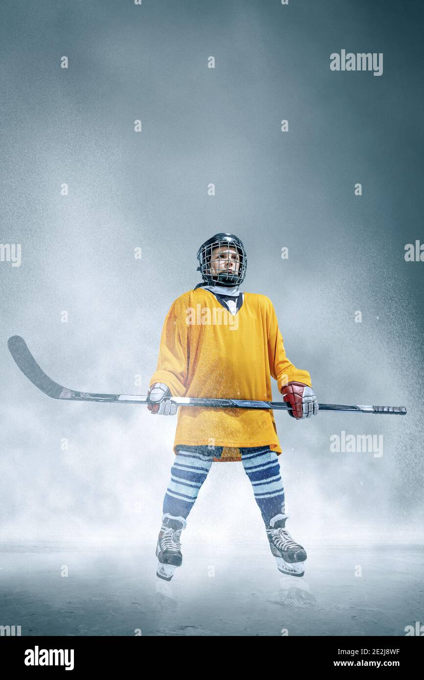 Campione. Piccolo giocatore di hockey con il bastone sul campo di ghiaccio e sfondo fumo. Attrezzature sportive e casco in azione. Concetto di sport, infanzia, movimento, movimento, azione. Foto Stock