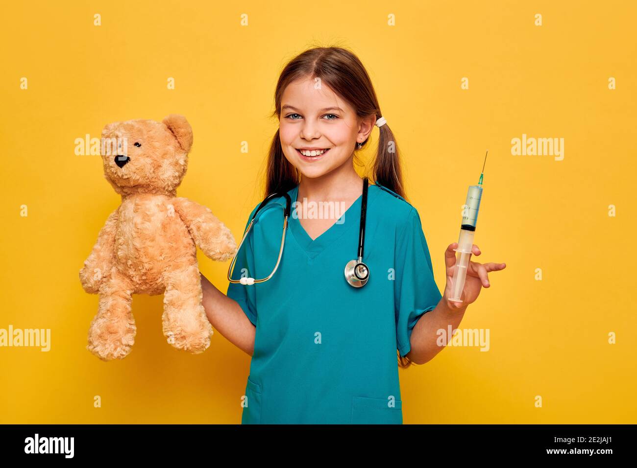 Positivo ragazza che va iniezione al suo paziente giocattolo orso. Bambino che gioca medico, infanzia, professione futura. Isolato su sfondo giallo Foto Stock