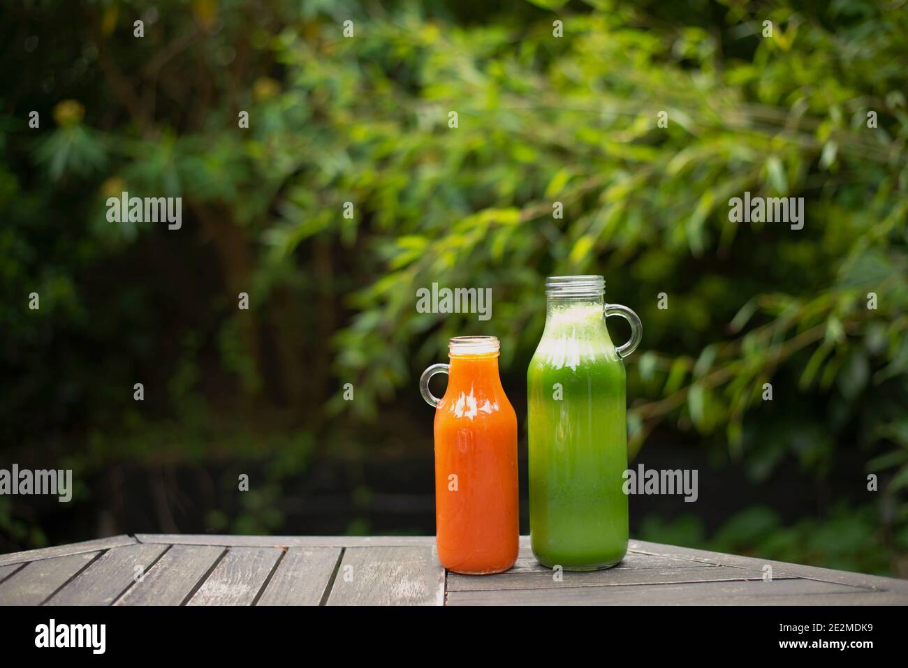 Succo fresco di verde e arancio su tavolo da giardino in legno. Foto di stile di vita all'aperto sano con sfondo verde lussureggiante. Concetto di disintossicazione pulita del succo vegano Foto Stock