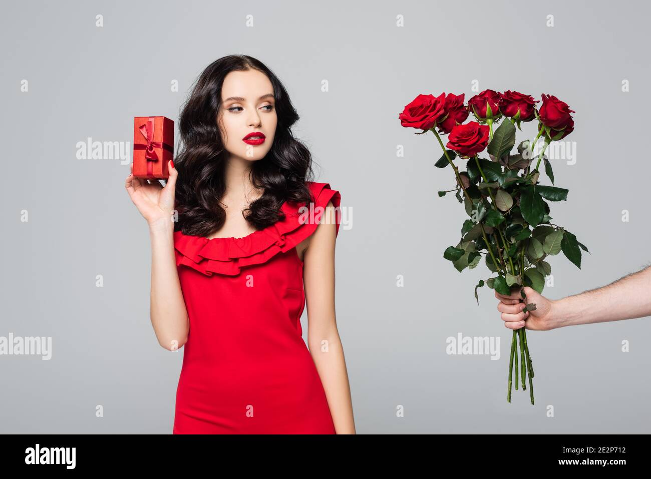 uomo che dà rose alla donna di brunette in vestito rosso che tiene confezione regalo confezionata isolata in grigio Foto Stock