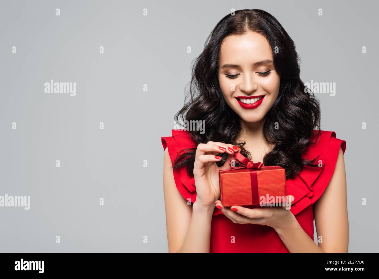donna allegra in abito con volant che tira il nastro sulla confezione regalo isolato su grigio Foto Stock