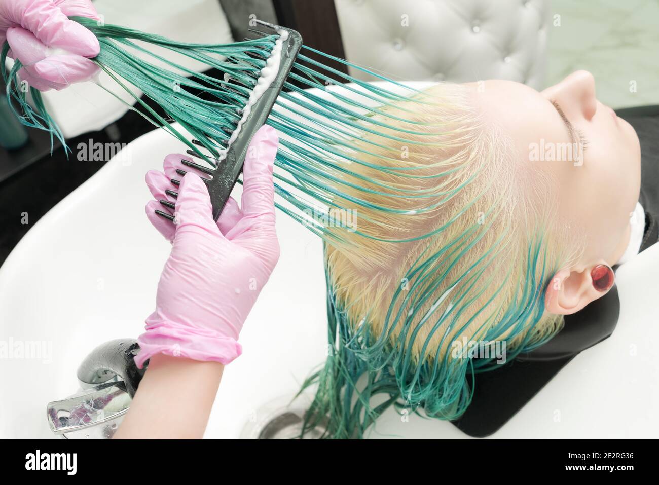 Parrucchiere in guanto protettivo che tiene in mano i capelli bagnati e pennellerà i capelli verdi lunghi e sbiancati del cliente adulto, mentre lavando la testa nella doccia Foto Stock