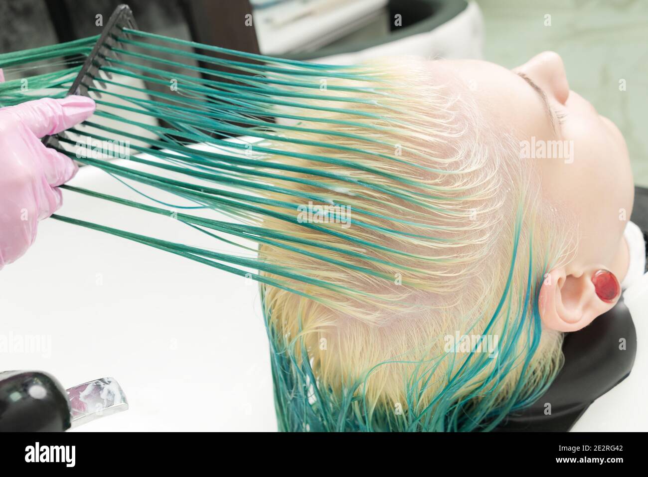 Stylist professionale in guanto protettivo tiene i capelli in mano e spazzola i capelli lunghi verdi e sbiancati del cliente, mentre lavano la testa nella doccia Foto Stock